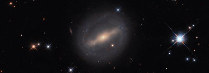 An almost circular galaxy is at the center of the image. It has a glowing bar stretching across its core; from the ends of the bar, thin spiral arms wrap around the galaxy to form a closed disk. The arms are fuzzy from the dust and stars they contain. The galaxy is on a black, mostly empty background. A few foreground stars with cross-shaped diffraction spikes can be seen, as well as some distant galaxies in the background.