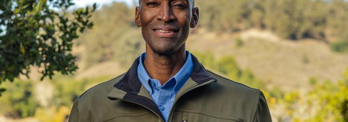 Rodney Martin, wearing an olive-colored NASA jacket and a blue dress shirt underneath, smiles at the camera. He is standing in the foreground with Lake Chabot Park in the background.