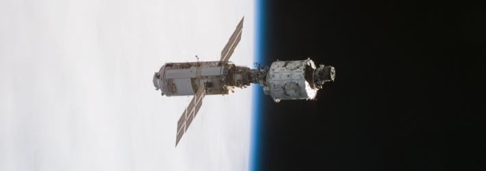 The first pieces of the International Space Station, the mated Russian-built Zarya (left) and U.S.-built Unity modules, are backdropped against the blackness of space and Earth's horizon. Zarya is cylinder-shaped and has solar panels that stick out from the body of the module.