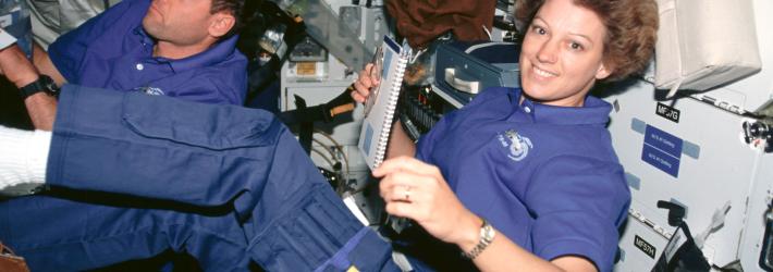 Two astronauts, one male (back) and one female (front) float inside the space shuttle. The woman in the front smiles at the camera while holding a spiral bound notebook in her right hand. She is wearing a blue polo shirt and blue pants with utility pockets on the thigh, as well as a gold watch on her left wrist. Her short brown hair floats upward.