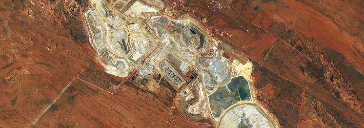 An overhead view of a mine in the dry outback of Australia. Most of the image is dominated by rusty red soil. Miners have excavated the soil in places, revealing an off-white color in these open pits. Some water is visible as well, in ponds.